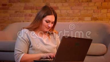 漂亮的加码模特坐在地板上，微笑着在笔记本电脑上打字，在舒适的家庭<strong>氛围</strong>中为工作<strong>感</strong>到高兴。